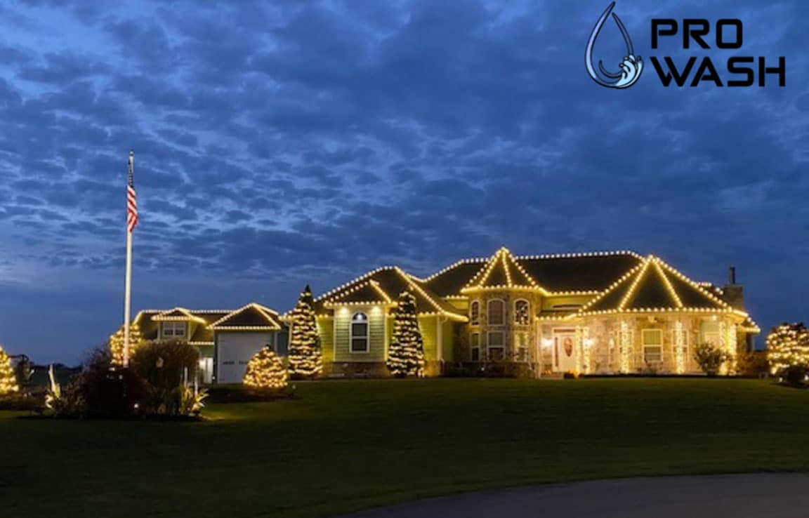 Large house in Farmington, Missouri, adorned with warm white holiday lighting outlining the roof and trees, creating a magical, inviting scene. An American flag waves in the yard. Pro Wash logo in the corner.