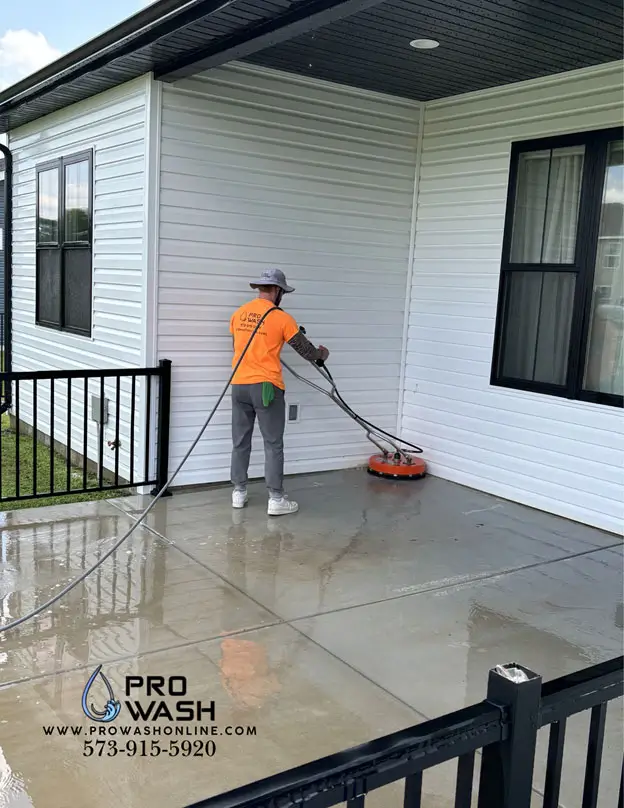 Person in orange shirt and hat using a circular surface cleaner to pressure-wash a wet concrete porch beside a white-sided house